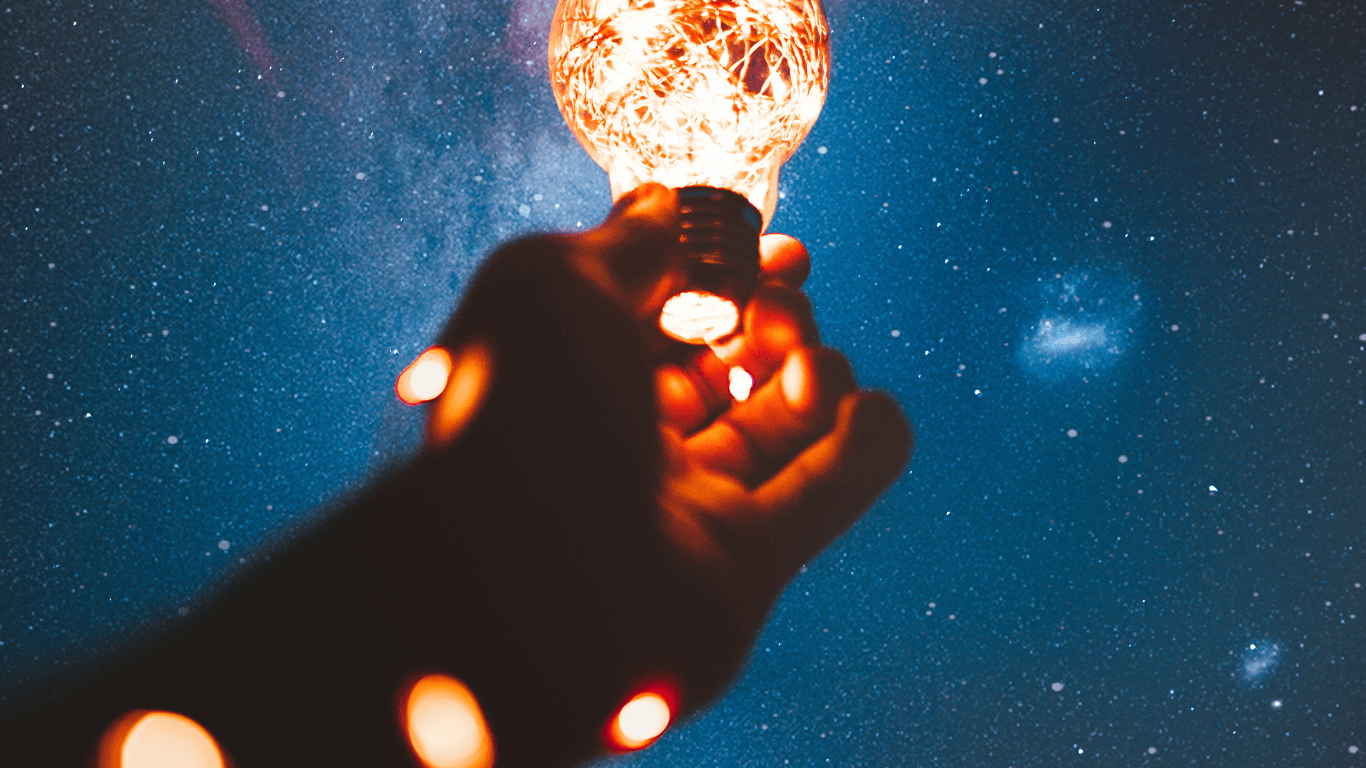 Hand holding a lightbulb against a starry night sky. 